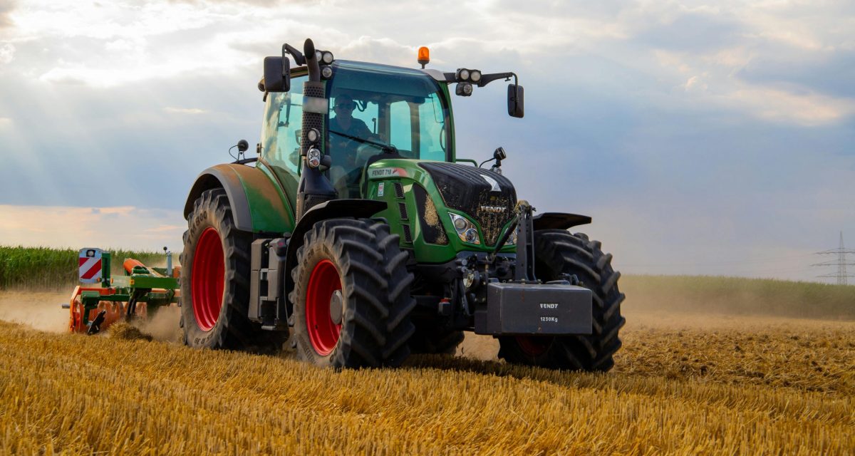 A powerful green tractor plowing a dusty wheat field under a cloudy summer sky in rural Germany.